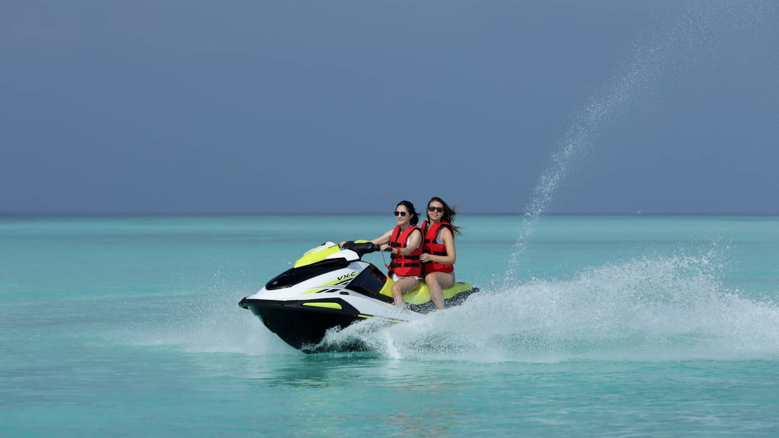 Wearing life jackets, two smiling jet-skiers ride through the clear ocean, water spurting up from beside and behind the boat.
