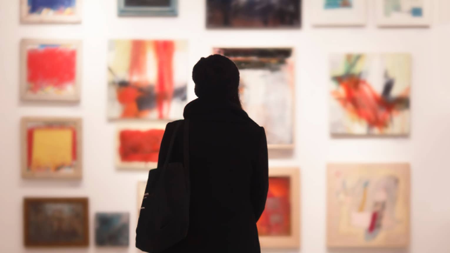 Silhouette of back of woman with hat bag looking at gallery wall with colourful art