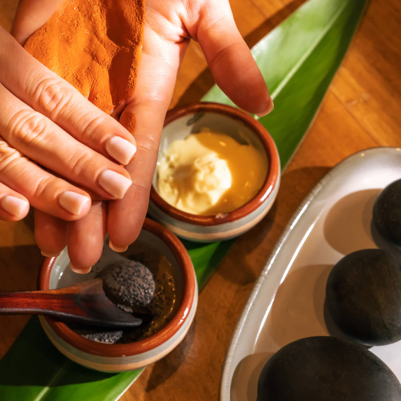 Pair of hands tests spa ingredients from two small stoneware bowls set on a fresh green leaf with three black stones placed on a dish nearby