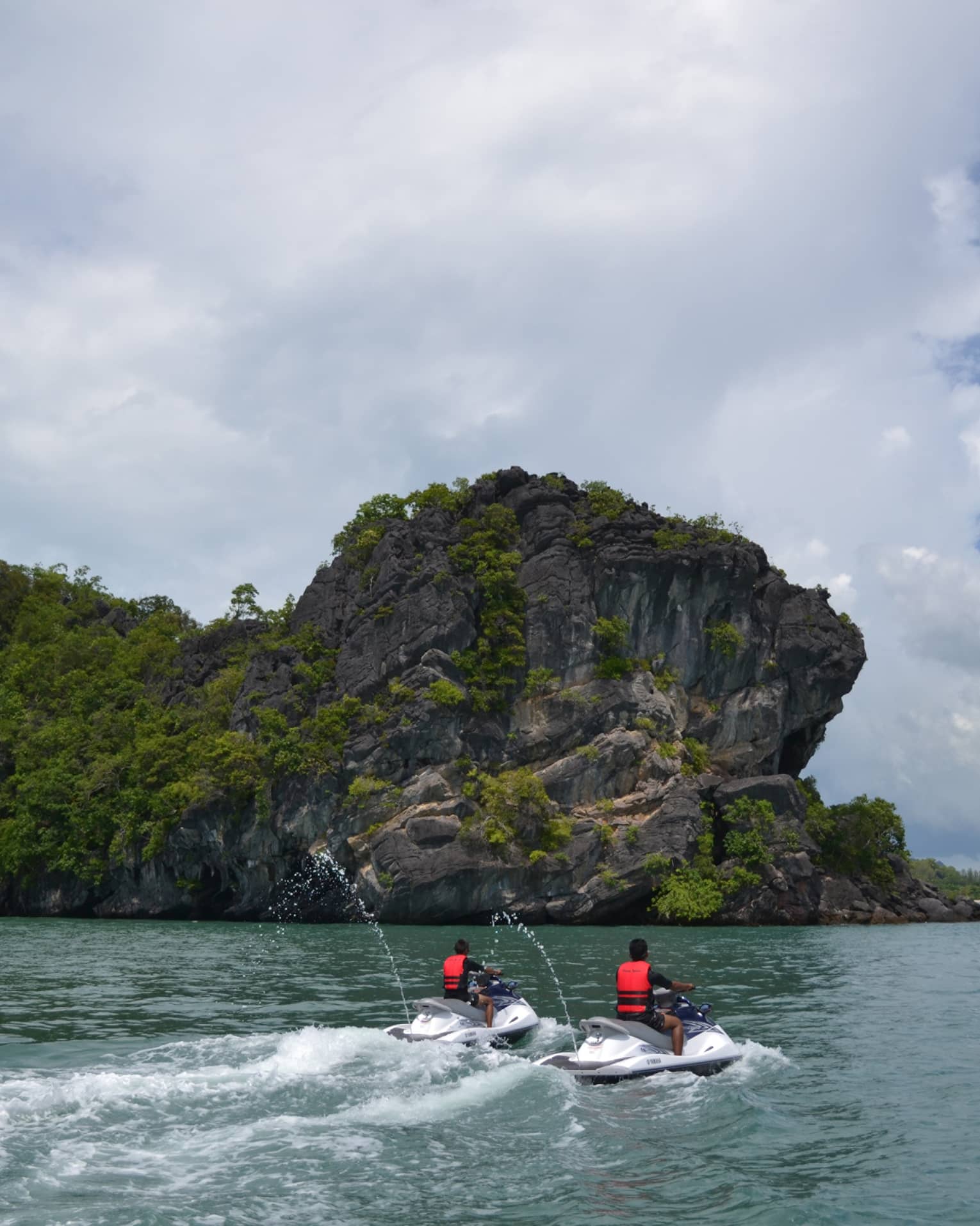 Two people wearing red life vests on jet skis ride on ocean under mountain